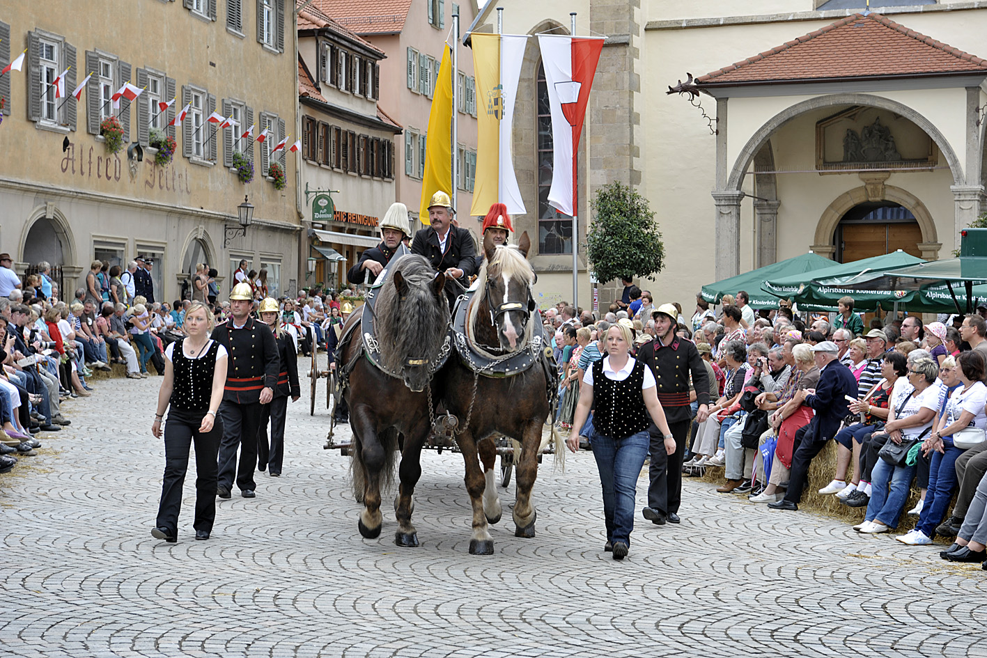 Eine Kutsche mit zwei straken Pferden. Die Kutsche wird von mehreren Personen in Uniform begleitet.