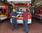 Zwei Männer stehen nebeneinander. Der eine gibt dem anderen einen Helm. 
