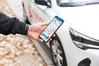 Ein Mann hält ein Smartphone hoch. Auf dem Display steht "Carsharing" im Hintergrund ist ein Auto zu sehen 