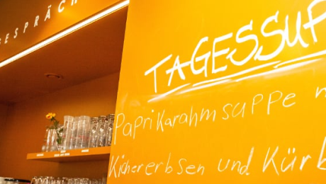 Orangefarbene Wand mit Regal im Hintergrund. Die Wand wurde beschriftet und Ausschnitte einer Speisekarte wurden darauf geschrieben.  Quelle: Café Stadtgespräch
