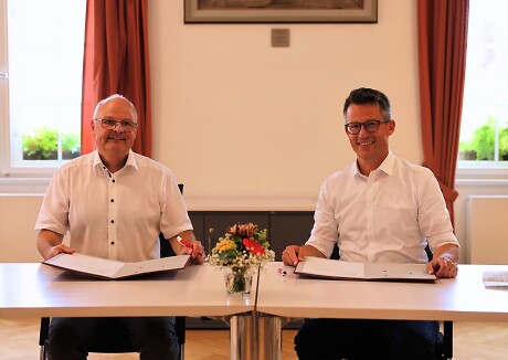 Dr. Hendrik Bednarz und Dr. Stephan Neu sitzen nebeneinander an einem Tisch. Beide lachen in die Kamera. Vor ihnen Liegt das Dokument der eben unterzeichneten Lernpartnerschaft.