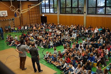 Turnhalle der Carl-Joseph-Leiprecht-Schule gefüllt mit Schülern auf dem Boden sitzend die zwei Referenten auf einer Bühne zuhören