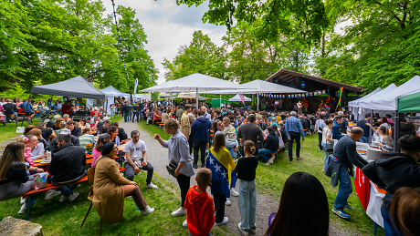 Viele Menschen stehen auf einer Wiese und feiern ein Fest. Manche sitzen an Tischen, manche holen sich essen an einem der Stände, die auf der Wiese verteilt sind.