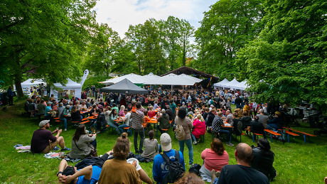 Fest der Nationen 2023 
Menschen sitzen auf einer Wiese und auf Bierbänken und schauen auf eine Bühne