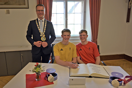 2 Junge Männer sitzen an einem Tisch und tragen sich in ein Buch ein. Der Oberbürgermeister steht links daneben.