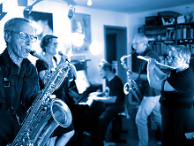 Die Band, links Saxofonist, dann Sängerin, Pianist, Tenorsaxofonist und Querflöte, alles in grau, blau Tönen gehalten