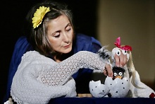 Eine Puppenspielerin animiert ein schwarzes Küken, das aus dem Ei schlüpft. Ihre andere Hand stellt die große, weiße Henne dar.