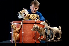 Verschiedene Tier-Puppen stehen um eine Trommel, auf der eine Tier-Puppe sitzt. Im Hintergrund der Puppenspieler.