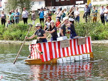 Ein Boot das als Pommes-Bude gestaltet ist, mit Kapitäninnen beim Kübelesrennen auf dem Neckar, am Ufer Zuschauer*innen